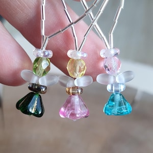 May include: Close-up of three beaded angel ornaments. Each angel features a different colored faceted bead body: green, yellow, pink, and blue. The wings are made of frosted glass, and the heads are clear beads. The angels are suspended by silver wire.