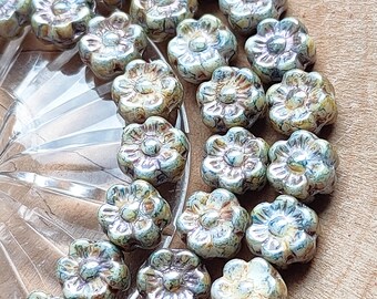 May include: A collection of flower-shaped beads in shades of green, brown, and cream. The beads are arranged on a clear, textured surface and a wooden background. The beads have a glossy finish and detailed petal design.