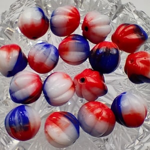 Puede incluir: Una colección de 15 cuentas de vidrio rojo, blanco y azul con un patrón de espiral. Las cuentas tienen forma de calabaza pequeña con la parte superior redondeada y la parte inferior ligeramente hundida.