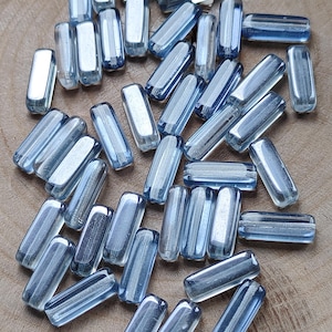 Könnte beinhalten: Eine Sammlung hellblauer, rechteckiger Glasperlen. Jede Perle hat abgerundete Kanten und ein durchscheinendes Aussehen. Die Perlen sind auf einer Holzoberfläche verstreut und zeigen ihre Farbe und Form. Ideal für die Schmuckherstellung oder das Basteln.