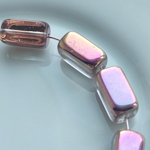 May include: A string of rectangular glass beads featuring a metallic, iridescent finish that shifts between shades of copper, pink, and purple. Each bead has rounded edges and is threaded onto a thin, silver-coloured wire, creating a smooth, reflective surface.