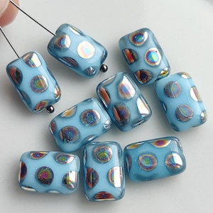 May include: A collection of rectangular glass beads in a light blue opaque finish. Each bead features multiple circular indentations with an iridescent, rainbow-like metallic sheen. Two of the beads are threaded onto thin metal pins for display.