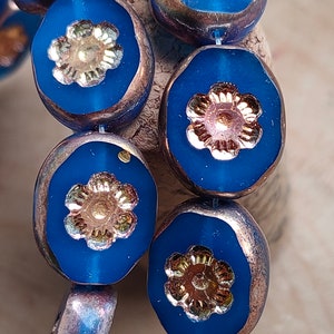 May include: A close-up of a strand of blue glass beads with gold and silver flower accents. The beads are iridescent and have a metallic finish.