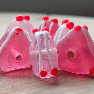 May include: A group of six pink glass beads with white and red accents. The beads are triangular in shape and have a hole in the centre for stringing.