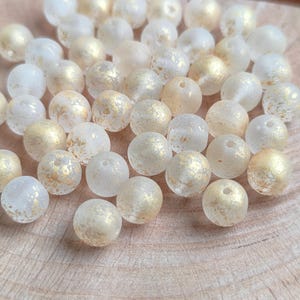 Puede incluir: Un conjunto de cuentas redondas y translúcidas con un aspecto esmerilado y detalles de pan de oro. Las cuentas están esparcidas sobre una superficie de madera clara, creando una estética texturizada y natural. Ideal para la creación de joyas.