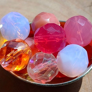 Può includere: Una collezione di perline sfaccettate in varie tonalità di rosa, rosso e arancione, presentate in un cucchiaio d'oro. Le perline hanno un design geometrico e sono realizzate in un materiale traslucido. Il cucchiaio è appoggiato su una superficie di legno.