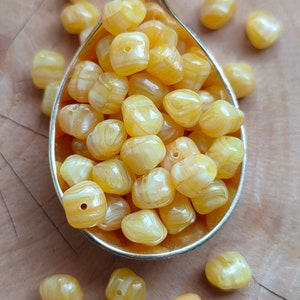 May include: A spoon filled with yellow and white swirled glass beads. The beads are square shaped with rounded edges. There are more beads scattered around the spoon on a wooden surface.
