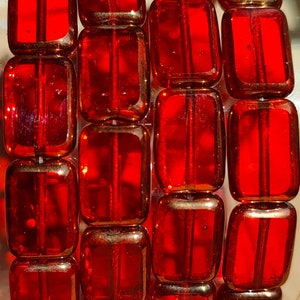 Op de afbeelding: Een close-up van een stapel rode glazen kralen met een gouden metallic afwerking. De kralen zijn rechthoekig van vorm en hebben een glad, glanzend oppervlak.