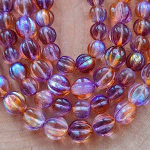 May include: A collection of round, ribbed glass beads featuring a translucent blend of purple and amber hues with an iridescent finish. The beads are arranged in multiple strands, showcasing a reflective surface that shifts between deep violet and warm orange tones.