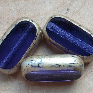 Könnte beinhalten: Drei violette Glasperlen mit goldenen Akzenten. Die Perlen sind oval geformt und haben eine flache Ober- und Unterseite.