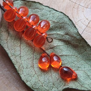 May include: A close-up of a decorative orange glass bead branch with teardrop-shaped beads. The beads are strung on a wire and arranged on a green leaf. The leaf rests on a wooden surface.