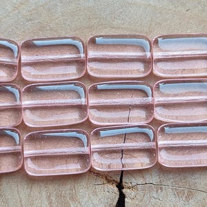 May include: A close-up shot of several translucent, rectangular beads in a soft pink colour. The beads are arranged in rows, showing their smooth, rounded edges and internal structure. The background is a light-coloured wooden surface.