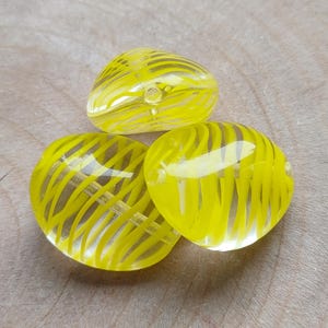 May include: Three translucent yellow glass beads with a striped design. The beads are oval and heart-shaped, with a hole for stringing. The beads are arranged on a wooden surface.
