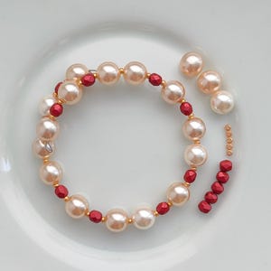 May include: A cream-coloured pearl bracelet with red beads interspersed. Several loose pearls and gold-coloured connecting rings are scattered around the bracelet on a white surface. A delicate piece of jewellery.