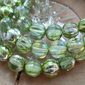 May include: A collection of round, translucent light green glass beads featuring a ribbed, melon-like texture. Each bead displays a subtle metallic sheen and vertical grooves, arranged in a cluster to highlight the reflective, glossy surface.