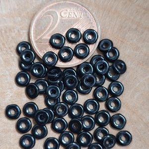 Könnte beinhalten: Eine Ansammlung kleiner, glänzender, schwarzer, ringförmiger Perlen auf einer Holzoberfläche, neben einer Zwei-Euro-Cent-Münze. Die Perlen haben ein Loch in der Mitte und reflektieren das Licht, wodurch ein glänzendes Aussehen entsteht.