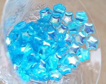 May include: A close-up of a bowl filled with iridescent blue star-shaped beads. The beads have a light blue color with a shimmery finish.