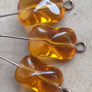 May include: Three amber glass beads with a distinctive, curved form. Each bead features a wire loop for threading and a wire through the centre. The beads have a glossy, transparent finish, suitable for jewellery making.