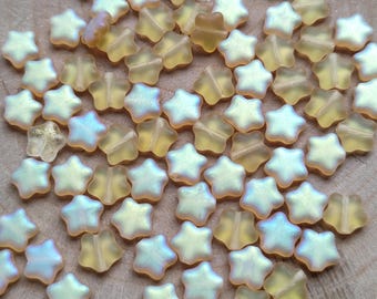 May include: A scattering of small, star-shaped beads. The beads have a translucent, iridescent appearance, with colours shifting from yellow to pale blue. The beads are arranged on a wooden surface, creating a textured and visually appealing display.