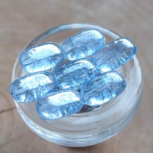 May include: A collection of seven light blue, oval-shaped glass beads inside a clear glass container. The beads have a textured surface and are arranged in a circular pattern. The glass container sits on a wooden surface.