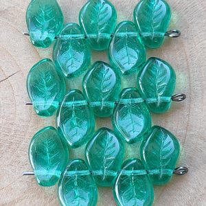 Può includere: Una collezione di perline trasparenti a forma di foglia di colore verde acqua. Ogni perlina presenta un motivo dettagliato di venature e viene infilata su un filo argentato. Le perline sono disposte in file, mostrando il loro design.
