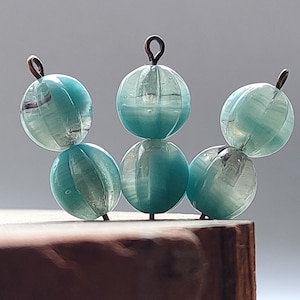 Puede incluir: Un grupo de seis cuentas de vidrio azul verdoso con un patrón estriado están enhebradas en un alambre delgado. Las cuentas están dispuestas en forma de semicírculo.