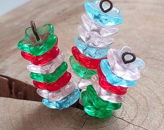 May include: Three stacks of colorful, translucent leaf-shaped beads. The beads are in shades of red, green, blue, and clear, with a small metal loop at the top of each stack. The beads are arranged on a wooden surface.