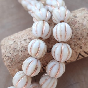 Puede incluir: Cuentas de vidrio blanco con textura acanalada y una fina franja dorada alrededor del centro. Las cuentas están enhebradas en un cordón y están sobre una superficie de corcho.
