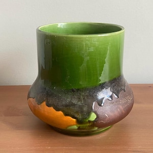 Peut inclure: Un vase en céramique avec une base large et arrondie et un dessus cylindrique. Le dessus est vert vif, passant à une base marron foncé et orange. Le vase a une finition brillante et repose sur une surface en bois.