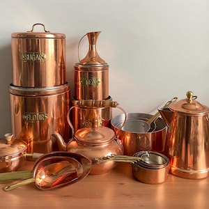 May include: A collection of polished copper kitchenware including two large canisters labeled SUGAR and FLOUR, a smaller TEA canister, a tea kettle with a wooden handle, a coffee pot, a saucepan, a set of measuring cups, and a large metal scoop.