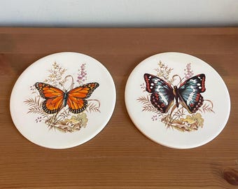 Pode incluir: Dois  copos de cerâmica branca com desenhos de borboletas. Um copo apresenta uma borboleta monarca com asas laranja e pretas, enquanto o outro copo apresenta uma borboleta azul e preta com manchas brancas.