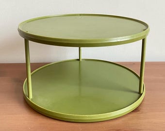 Pode incluir: Uma bandeja de servir de dois níveis, redonda, verde-oliva. A bandeja tem um design simples com uma parte superior e inferior planas, conectadas por quatro suportes verticais. A bandeja é feita de um material sólido e fica em uma superfície de madeira.