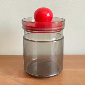 Peut inclure: Un bocal en verre transparent avec un couvercle rouge et une poignée rouge en forme de boule. Le bocal a une forme cylindrique et est vide. Le couvercle est un disque plat avec un bord surélevé. Le bocal est posé sur une surface en bois.