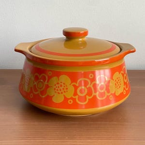 Peut inclure: Un plat à casserole vintage en céramique avec un couvercle. Le plat est orange avec un motif floral jaune et un bord marron. Le couvercle a un bouton marron et des rayures orange. Le plat a deux poignées.