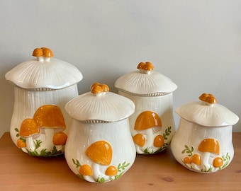 Pode incluir: Conjunto de quatro potes de cerâmica branca com desenhos de cogumelos. Os potes são decorados com cogumelos laranja e folhas verdes.