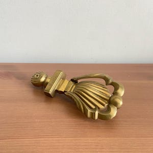 May include: A brass door knocker with a shell-shaped design and a handle. The knocker has a rectangular base with the word "Carter" engraved on it. The knocker is lying on a wooden surface.