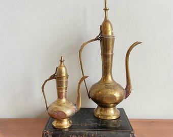 Pode incluir: Duas jarras antigas de latão com desenhos ornamentados. A jarra maior tem um bico longo e um topo pontiagudo. A jarra menor tem um corpo arredondado e uma tampa articulada. Ambas estão em um livro vintage.