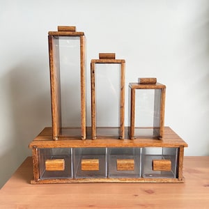 May include: Set of four wooden storage canisters with clear glass lids. The canisters are arranged in descending size order and are sitting on a wooden shelf with four drawers.