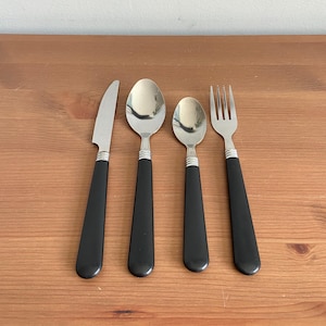 May include: A set of four stainless steel utensils with black handles. The set includes a knife, a large spoon, a small spoon, and a fork. The utensils are arranged on a wooden surface.