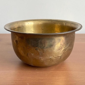 May include: A vintage brass bowl with a rounded shape and a wide rim. The bowl has an aged appearance and features delicate floral engravings around the exterior. The interior of the bowl is smooth and reflective.