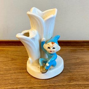May include: A vintage ceramic vase in the shape of a white flower with a blue elf figurine. The elf is wearing a blue outfit and hat, with a red mouth and orange hair. The vase is on a white base.