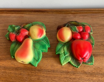 May include: Two three-dimensional wall plaques featuring molded fruit designs. The left plaque displays a pear, a peach, and strawberries nestled in green leaves. The right plaque features a large red apple, a peach, and cherries with green leaves.