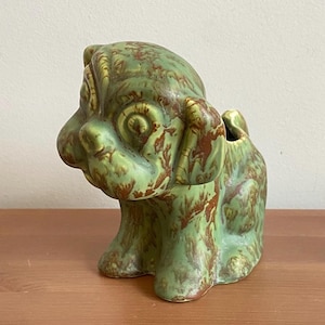 May include: Green ceramic dog planter with brown marbling. The dog is sitting with its head tilted to the side and its ears perked up.