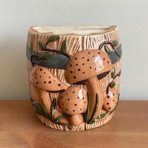 May include: A ceramic wall planter shaped like a wooden barrel with a brown, green, and white glaze. The planter features three mushrooms with brown and white spots.