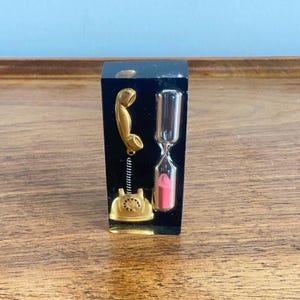 May include: A rectangular black resin block containing a miniature gold-coloured rotary telephone with a coiled cord and a small hourglass filled with pink sand. The objects are embedded within the clear, dark material, creating a solid decorative piece.