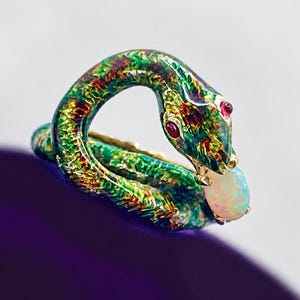 May include: A snake-shaped ring featuring a textured green and gold finish with small red gemstone eyes. The serpent's head is curved, holding a smooth, oval-shaped iridescent white opal in its mouth, creating a continuous circular band design.