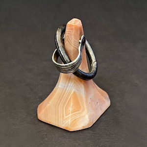 May include: A silver ring with a black metal band wrapped around it. The ring is displayed on a pink and white agate stand.