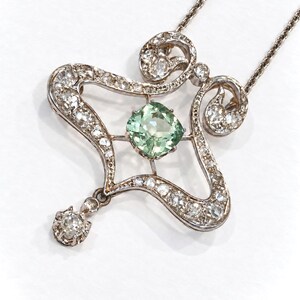 May include: A silver-toned pendant necklace featuring a central cushion-cut light green gemstone surrounded by a decorative openwork frame set with numerous small clear stones. A single clear stone drop hangs from the bottom of the frame.