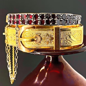 May include: Two antique bracelets. The top bracelet is silver with small, dark red gemstones. The bottom bracelet is gold-toned with floral and scrollwork designs. A gold chain hangs from the side.