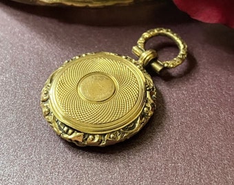 May include: A gold-colored, round antique locket with intricate detailing and a decorative loop. The locket is next to a matching compact and a dark red flower, all on a purple surface. The locket's design features a textured pattern.